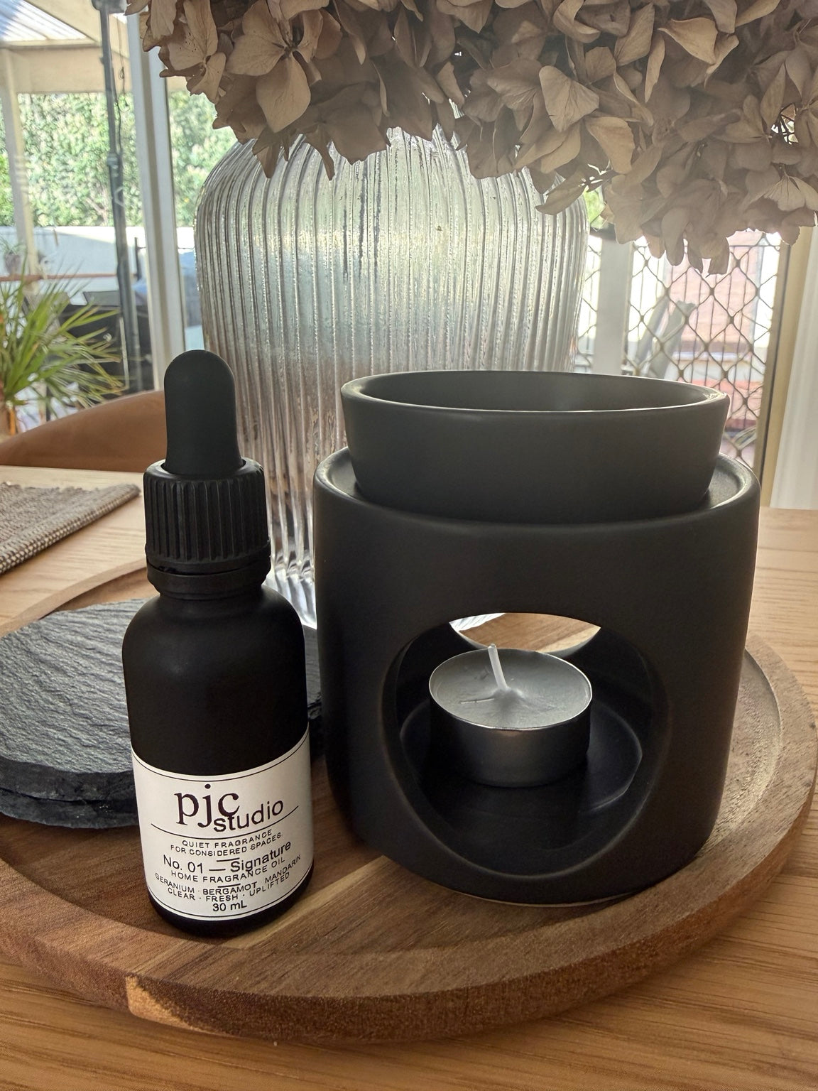 Close view of the larger matte black oil burner, its removable upper bowl and tealight chamber, beside No. 01 Signature Home Fragrance Oil. The duo set includes two fragrance bottles.