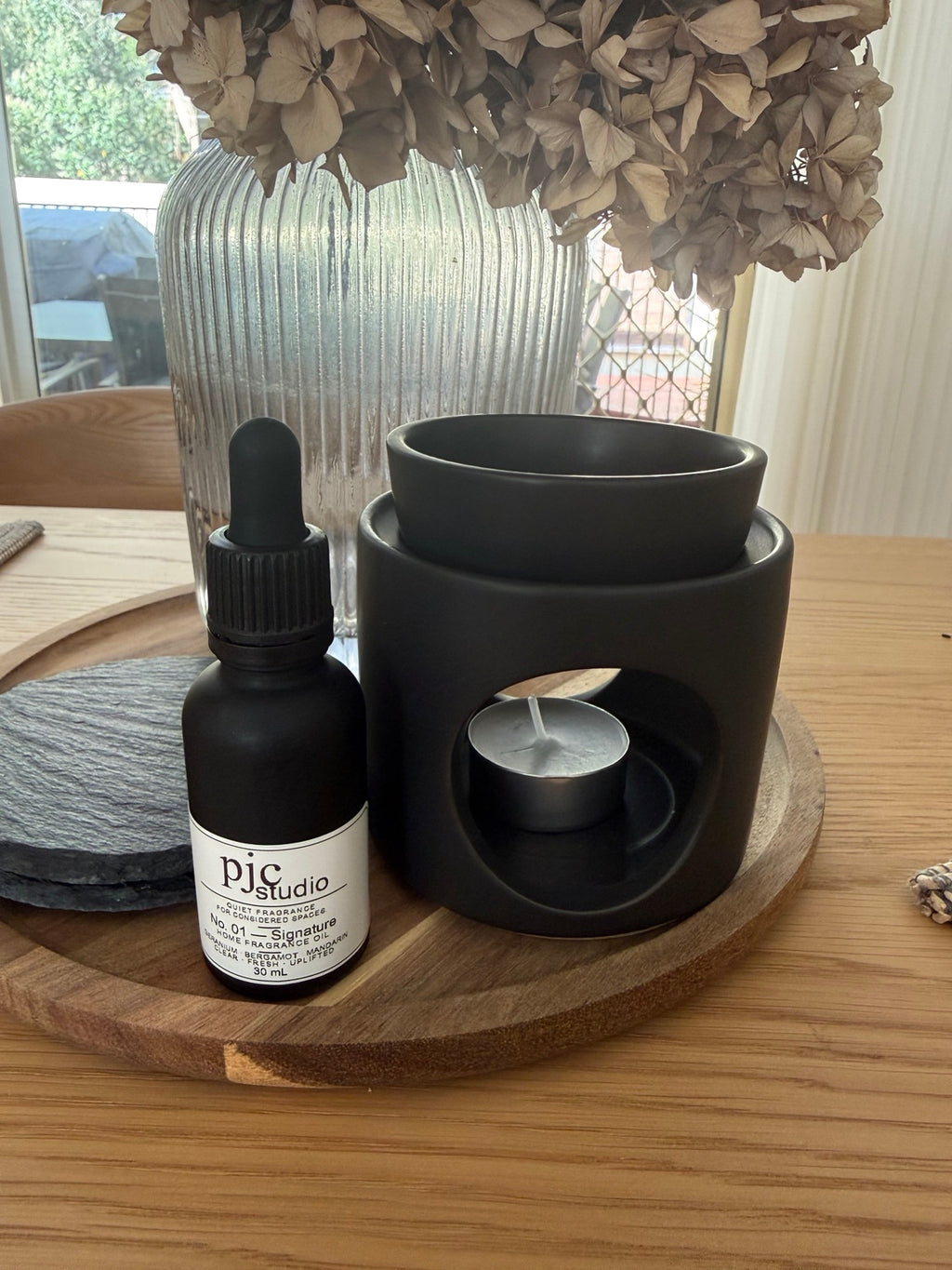 Larger matte black ceramic oil burner with removable bowl, styled beside a 30 ml bottle of pjc studio No. 01 Signature Home Fragrance Oil on a wooden tray.